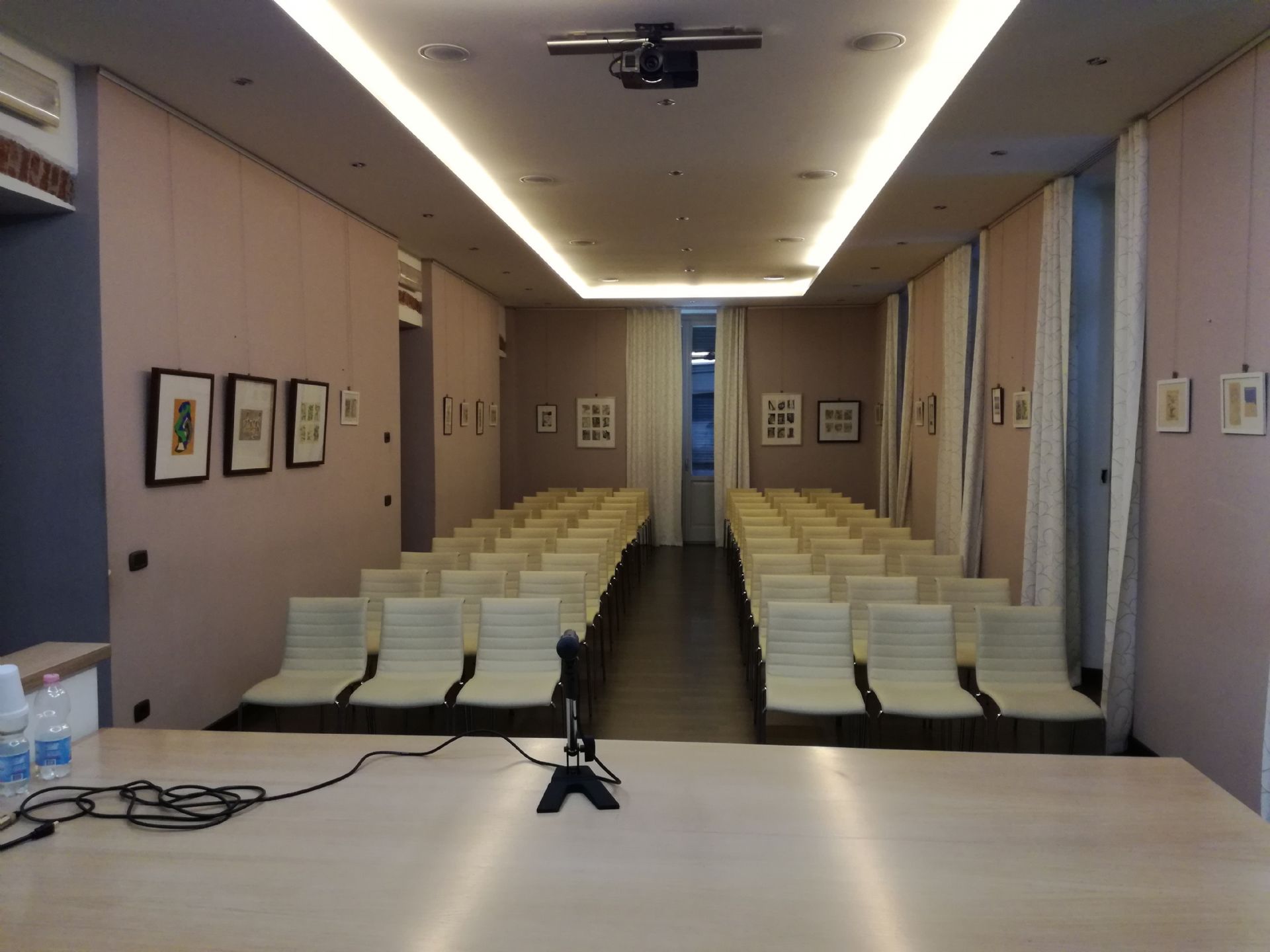 Sala polivalente con  sedie e tavolo con microfono in primo piano. Esposizione di quadri sulle pareti. Videoproiettore al soffitto e luci soffuse
