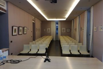 Sala polivalente con  sedie e tavolo con microfono in primo piano. Esposizione di quadri sulle pareti. Videoproiettore al soffitto e luci soffuse